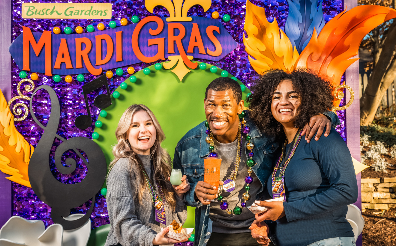 Three people holding drinks in front of a Mardi Gras sign